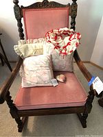 Front view of the rocking chair with plush pink velvet seat and backrest, displaying four assorted decorative throw pillows with lace and red ribbon accents on the seat.