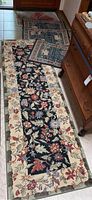 Photo showing one long floral runner rug with black center and cream floral border, alongside four small area rugs with patchwork and geometric design.