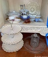 Three-tiered ceramic serving stand with scalloped edges and gold trim. Glass biscuit jar with lid. Various tea cups, saucers, and serving bowls in the background on a carved wooden surface.