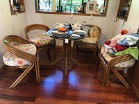 Full set of round rattan table with glass top and four matching chairs with floral cushions in a room with hardwood floors.