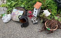 Photo showing metal watering can, bird house, hummingbird feeder, swan statue, and pots with plants and soil outdoors on pavement.