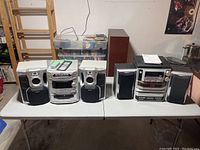 Wide shot of both stereo systems, showing all components including speakers, main units, and remote, on a white table in a storage or basement space.