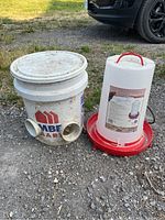 Photo showing a homemade feeder made from a five-gallon bucket with circular openings and a white and red heated waterer with handle and power cord.