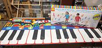 Gigantic step & play piano floor mat with oversized piano keys, colorful border, and original box behind it showing children playing on the toy.