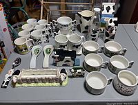 Photo showing multiple ceramic mugs with black Holstein cow patterns and cow-shaped handles, soup bowls, spoons, napkin holders, and a rectangular container with cow figurines at each end, all with a Holstein theme.