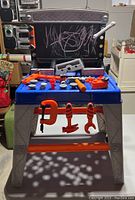 Front view of the kids tool bench toy with assorted orange and gray plastic tools on the blue work surface and hanging from the bench. Chalkboard back panel with chalk scribbles visible.