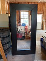Full view of large black framed mirror standing vertically with visible surroundings including a stack of tires and household items.
