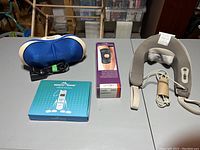 Wide view of all items included in the lot on a grey table; shows blue BackPlus massager, knee brace box, Easy@Home TENS device box, and grey neck massager with wires.
