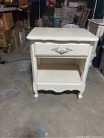 Front view of the single white side table with drawer and open shelf, showing ornamental hardware and carved legs.