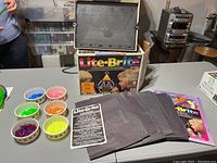 Lite Brite toy set displayed with light board upright on original box, several black design pages spread out, colorful pegs in separate bowls, and instruction sheet visible, showing the lot contents.