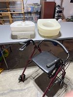 Front angle of the Bios walker with seat and basket, two raised toilet seats on table behind walker.