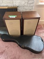 Pair of vintage wooden Advent speakers with beige fabric covers and acoustic guitar in black hard case on carpet in front of fireplace.