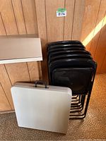 Six black folding chairs stacked against wall next to white folding table.