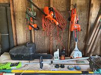 Several hedge trimmer and chainsaw attachments hanging on shed wall above table; black scabbard case at left
