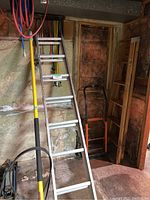 Three ladders leaning against garage wall: aluminum extension ladder, wooden step ladder, and metal dolly/hand truck; pressure washer gun partially visible.