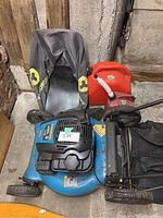 Full view of the blue Yardworks lawn mower with black engine cover, red gas can, and black grass collection bag on concrete floor in garage.