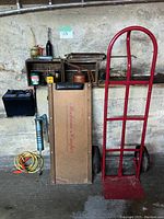 Photo showing mechanic's creeper, hand dolly, car battery, and small workshop items on shelf against concrete wall