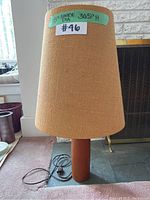 Full view of the lamp showing tapered teak wood base, fabric shade, electrical cord and plug on carpeted floor near a fireplace.
