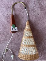 Wall sconce laid flat showing wood triangular mount, brass arm, and conical wicker shade on carpet