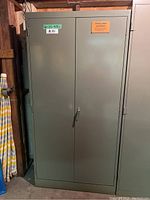 Front view of gray metal storage cabinet with two doors closed, showing handle and stickers on top right and left describing size and lot number