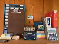 Photo showing a blue storage drawer organizer with small compartments, brown clipboard, ink cartridge box, basket with office tools, Dymo label maker, colorful index cards, and address label boxes.