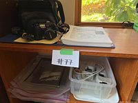 Overview of camera and accessories on wooden shelf including camera manual binder underneath and storage bin with cables and charger.