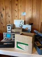 Overall view of the lot showing oscilloscope, multimeter, headphones, and boxed accessories on table against wooden wall.