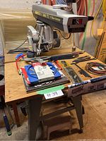 Full view of the DeWalt 770 Deluxe Powershop radial arm saw mounted on a metal stand with wooden top, as well as various woodworking accessories and tools placed on the table around the saw.