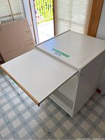 White kitchen trolley showing pull-out extension panel extended from one side, with light wood edge trim, against a window with blinds.