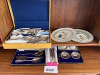 Wood shelf display of two flatware cases with assorted forks, spoons, knives, two decorative plates on upper shelf, a box of table knives and two salt cellars on lower shelf