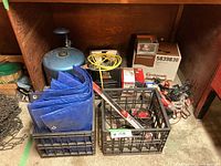 Items under wooden shelf including crates with tarp, jumper cables, and various small items