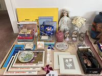 Lot overview showing art supplies, scrapbooks, jewelry, porcelain doll, and decorative items arranged on floor.