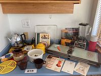 Shows assortment of vintage kitchenware items including kettle, cups, hot trays, blender, and cookbooks arranged on blue countertop.