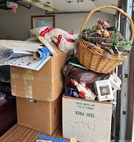 Stacked cardboard boxes containing various Christmas decoration items including a wreath basket on top, tree skirts, and wrapping paper.