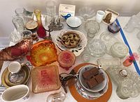 Lot of vintage glassware and china on a table featuring Amberina carnival glass candy dishes, cut glass ashtrays, ceramic bowls, plastic bird salt and pepper shakers, brass candle holders, shot glasses, and a boxed figurine.