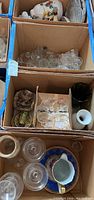 Top view of multiple boxes containing clear glass margarita glasses and stemware, candy dishes, plates, and other assorted glass pieces along with a floral gold-accented porcelain jug