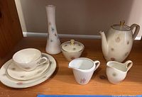 Full set of ceramic tea set pieces including teapot, creamer, sugar bowl with lid, cup and saucer, two plates and a vase displayed on wooden shelf.