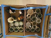 Top-down view of two boxes containing a mix of 18 mugs and 2 decanters, showing handles and some shapes like the stein and pourer.
