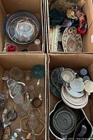Four cardboard boxes filled with various household items including floral teacups, glassware, scarves, and plates