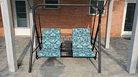 Front view of the metal outdoor swing with two blue patterned cushions, black metal frame, and central decorative metalwork between seats.