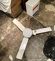 Entire ceiling fan laid flat showing all three white blades and central brushed metal housing with dome light cover. Remote control placed on one of the blades.