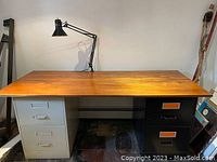 Desk setup showing the wooden top, two filing cabinets (light gray and black) underneath, and the desk lamp on the left side.