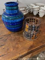 Blue glazed ceramic vase and clear polka dot glass shown on wooden surface with background of white cups and silver tureen.