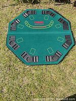 Full view of octagonal poker table top laid flat on grass, showing all nine player positions with molded black plastic chip trays and drink holders, and green felt surface with casino design.
