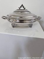 Full view of the closed silver plated round serving dish with lid and decorative handle on top, showing the ornate feet and side handles.