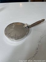 Closed lid view of the silver silent butler with wooden handle, showing etched design on top and turned wood handle attached to silver body.
