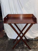 Front view of wood serving tray on folding stand, showing tray with raised sides and cutout handles, and cross-legged folding base.
