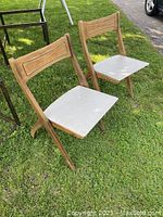 Two wood folding chairs with light colored vinyl seats placed outdoors on grass, showing front and side angles.