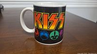Side view of white ceramic coffee mug with large KISS band logo and colorful band member icons on black background.