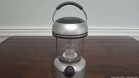 Full view of a portable silver lantern with round handle on top, transparent plastic middle section, and a control knob at base.